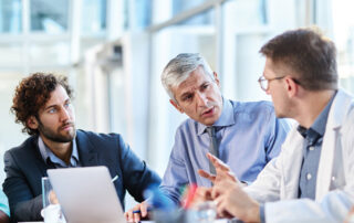 Group of healthcare professionals and business colleagues sitting at a table in a bright office, discussing something together.