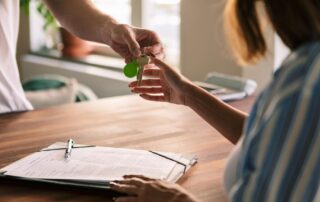 Staying Ahead Of The Curve: The Rental Market Trends That Will Define 2026 5 Person handing over house keys to another person across a table with signed contract paperwork.
