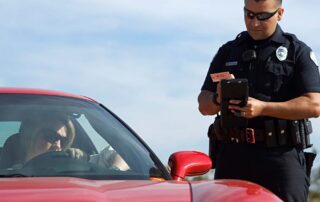 Driving With An Expired License In 2025: What Professionals Need To Know Before Hitting The Road 3 Police officer standing beside a red sports car, reviewing a driver’s license and issuing a traffic citation to the driver.
