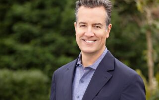 Smiling man in a navy blazer posing outdoors with greenery in the background