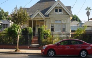 Red sedan parked in front of a fenced victorian-style house with a small front yard.