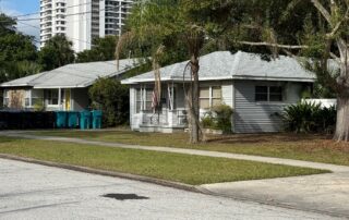 Single-story gray bungalow-style house with front porch, lawn, and large tree in a suburban neighborhood.