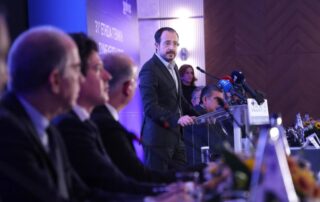 Cyprus Unveils Aggressive Housing Reforms Aimed At Faster Development And Greater Affordability 7 Man at podium speaking at formal conference with panel of people seated at a long table