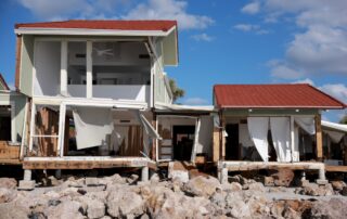 Damaged coastal house with red roof and exposed interior after a storm, sitting on broken pilings above rocks.