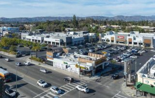 Aerial view of a busy suburban shopping center with large parking lot, grocery store, restaurants, and cars at a main street intersection.