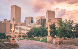 Downtown city skyline at sunset behind a tree‑lined park with stone columns, steps, and people walking.