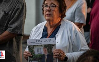 A woman at a public gathering holds a printed photo sign that reads “free jose” in support of a man pictured in front of bice ristorante palm beach.