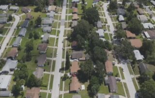 Aerial view of a suburban neighborhood with single‑family homes, green lawns, trees, and straight residential streets laid out in a grid pattern.