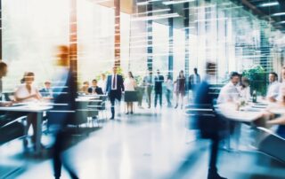 Busy modern office lobby with blurred motion of people walking and meeting at tables in a bright glass-walled workspace.