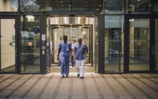 Hospital staff in blue scrubs walking out of a modern hospital entrance through glass revolving doors.