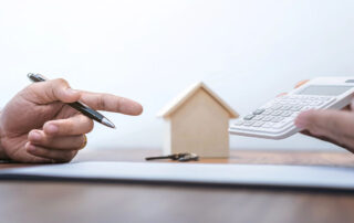 Two people discussing home financing with a small wooden house model, a set of keys, and a calculator on a desk.