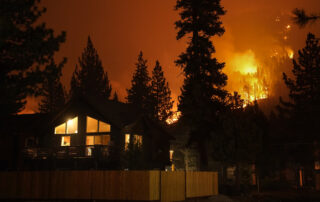 Nighttime view of a cabin-style house with lights on as a large forest wildfire burns on the hillside and in the trees behind it.