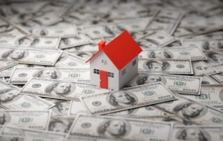 Small model house with a red roof sitting on top of scattered us hundred-dollar bills