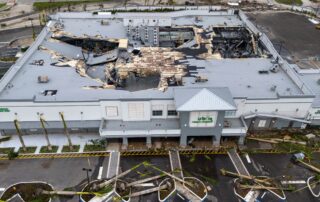 Aerial view of a supermarket building with a partially collapsed roof and surrounding storm damage after a hurricane.