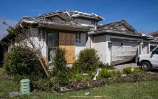 Hurricane-damaged suburban home with boarded windows, collapsed garage door, and debris scattered across the front yard.