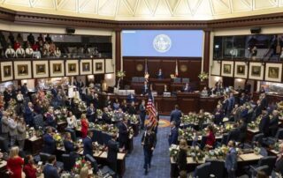 Members of a legislative body stand at their desks with floral arrangements as a color guard carrying the american flag walks down the center aisle in a large government chamber.