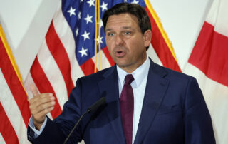 Man in a dark blue suit speaking at a podium in front of u. S. And florida flags