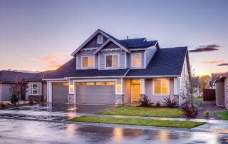 Modern two-story suburban house with three-car garage at sunset, lights glowing inside and wet driveway in front