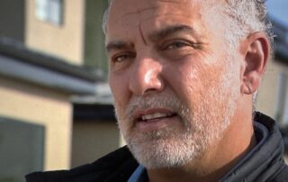 Close-up of a middle-aged man with gray hair and beard standing outside near a modern house.