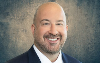 Smiling bald man in a suit and tie posing for a professional headshot against a textured blue background.
