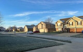 Suburban residential street with two-story houses, driveways, and cars on a clear blue morning