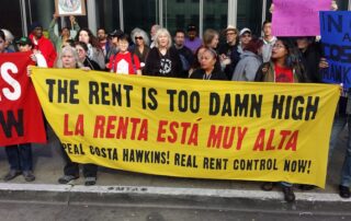 A diverse group of protesters hold a large yellow banner reading “the rent is too damn high / la renta está muy alta – repeal costa hawkins! Real rent control now! ” during a housing rights demonstration.
