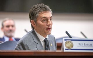 Person in a gray suit speaking into a microphone at a formal meeting, with a nameplate reading “rep. Basabe” in front of them.