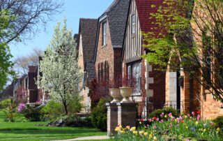 Row of brick houses with gabled roofs, flowering trees, green lawns, and colorful spring tulips along a quiet residential street.