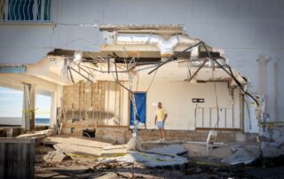 A person stands inside a beachfront building with its outer wall torn away, exposing severe structural damage and debris after a storm.