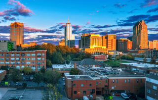 Sunset skyline view of a modern city with tall office buildings and apartments above a mix of trees and brick structures in the foreground.
