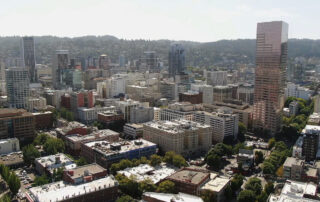 Aerial view of a modern downtown cityscape with tall office buildings, mid-rise apartments, and tree-lined streets on a clear day.