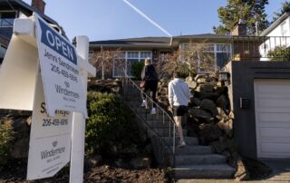 Prospective homebuyers walk up the front steps toward a house during an open house, with a real estate open sign visible in the foreground.