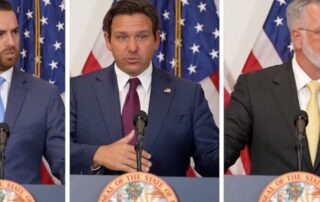 Three suited men stand at separate podiums with microphones in front of u. S. And state flags, speaking at a formal press conference.