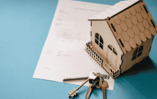 Wooden model house and metal keys resting on top of a real estate contract on a blue background.