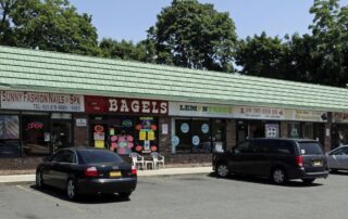 Small suburban strip mall with green roof, brick storefronts, and several parked cars in front of nail salon, bagel shop, hair salon, chinese restaurant, pet grooming, and pizza place.