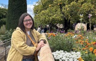 Smiling person in a yellow jacket standing beside a stone railing in a colorful flower garden with trees in the background.