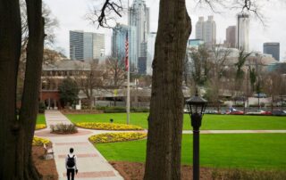 Student walking through a university campus park with flower beds and a city skyline in the background.