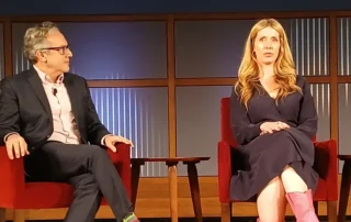 Two speakers seated on stage in red armchairs during a panel discussion, with wooden paneling and vertical glass strips in the background.