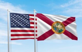 Two flags flying on flagpoles against a blue sky, the united states flag in the foreground and the florida state flag in the background.