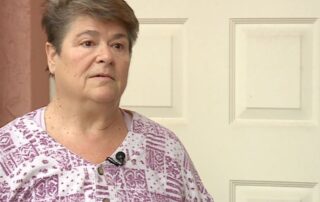 Older woman standing in front of a closed door, wearing a patterned shirt and a small clip-on microphone.