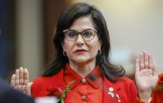 Woman in a red suit with a rose corsage raises her right hand during a formal ceremony.