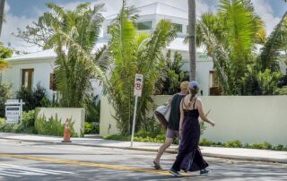 Couple walking barefoot across a sunny street in front of a modern tropical house with palm trees.