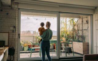 Mother holding toddler while looking out at balcony garden through large glass doors