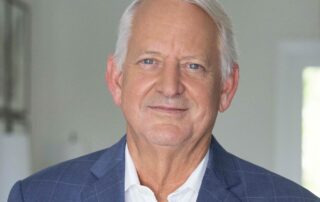 Portrait of an older man in a blue suit jacket and white shirt, smiling slightly in an indoor setting.