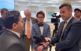 Group of reporters interviewing a man in a suit inside a government or office building, with microphones and a video camera recording the conversation.