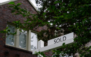 Sold sign in front of a brick house partially hidden by green tree branches.